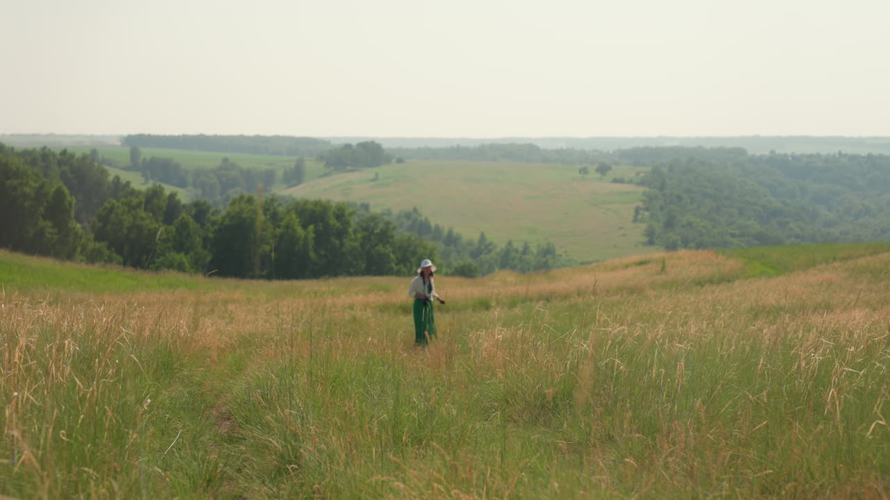 Rear view of woman in white hat and green skirt walking along grassy path through countryside, pausing to turn around amid rolling hills, distant forest, and golden summer fields