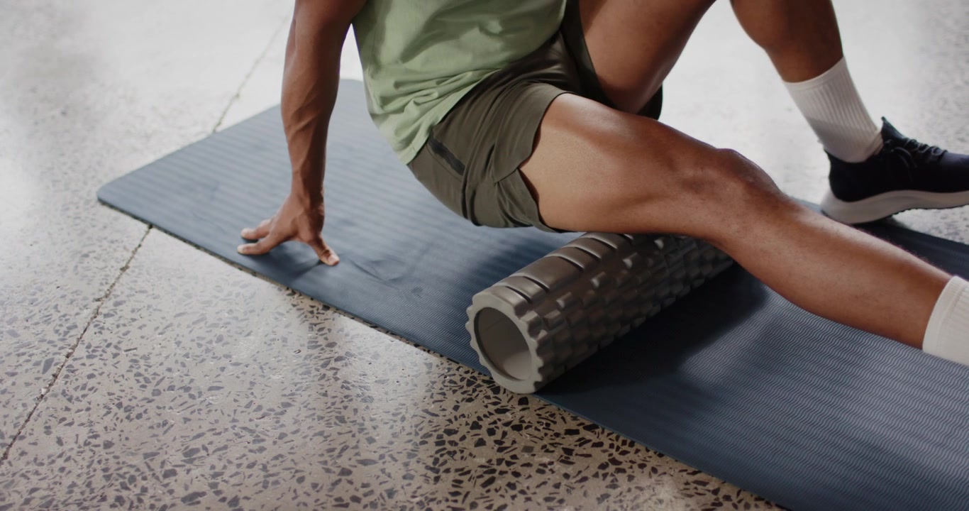African American man placing foam roller under right thigh, rolling to relieve tension on blue mat