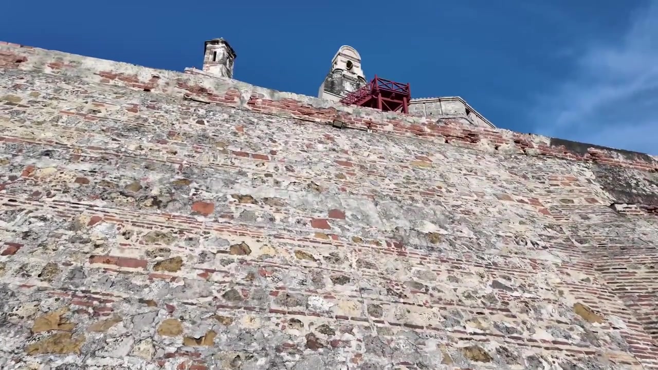 Una drammatica prospettiva dal basso che guarda verso le immense, storiche mura di fortificazione in pietra e mattoni del castello di San Felipe sotto un cielo blu intenso.