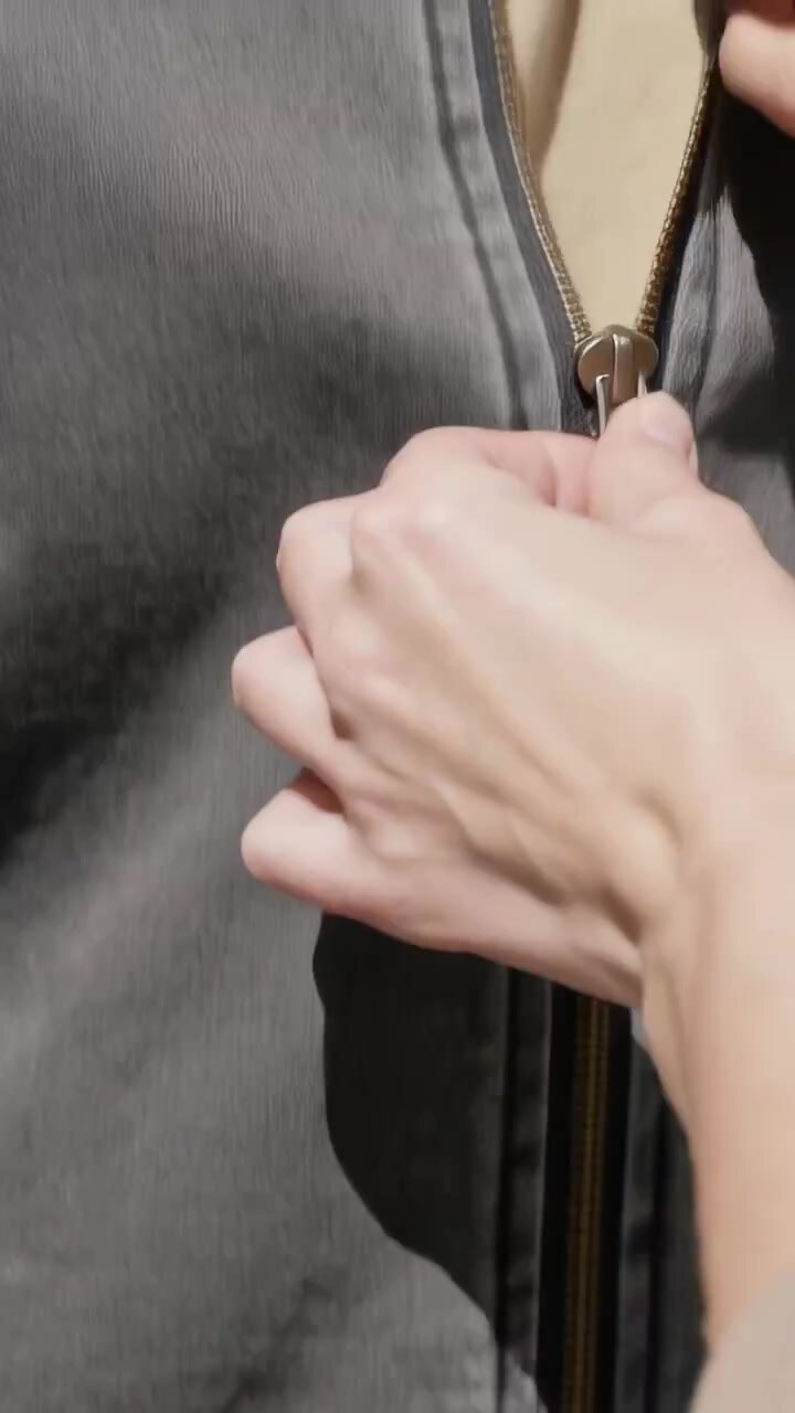 Vertical video: Gripping metal slider, right hand zipping gray jacket at home, showing beige shirt