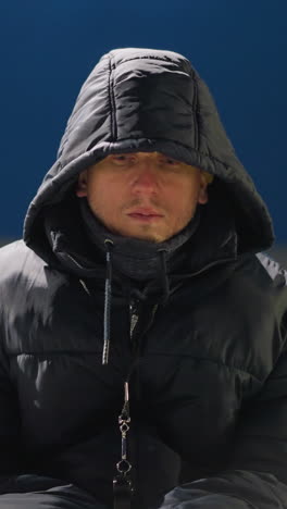un hombre con un traje negro y una chaqueta con capucha se sienta solo con las manos entre las piernas, pareciendo sobrio, está en un estadio con un fondo débilmente iluminado
