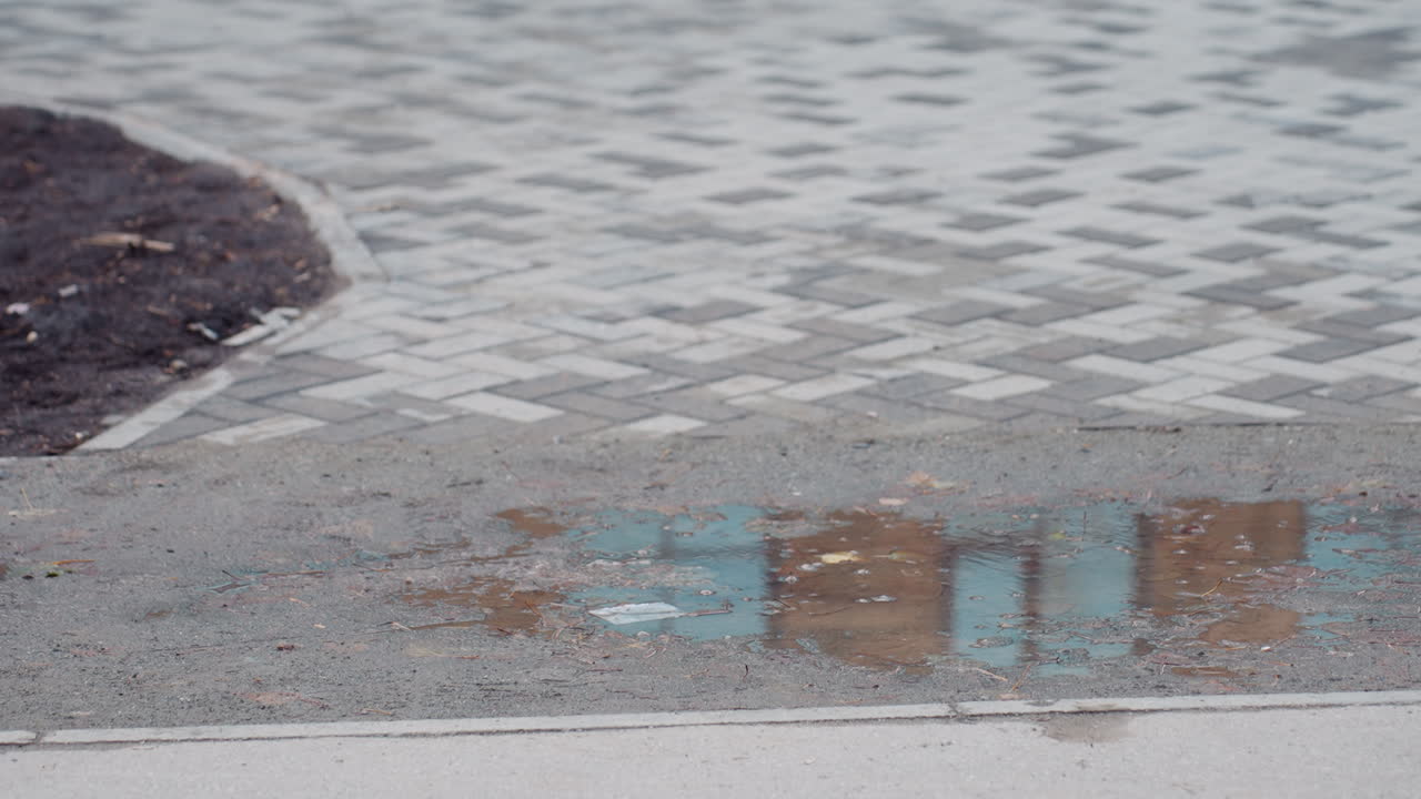 Wet tiled walkway with small puddle reflecting glass facade of nearby building as inline skater glides past, capturing motion, texture, and urban outdoor atmosphere in subtle overcast lighting