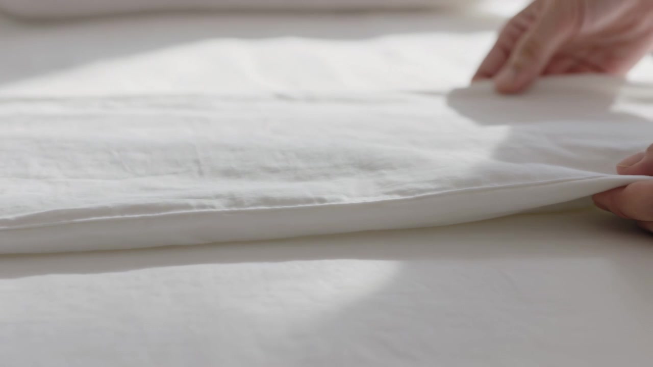 Smoothing bare hands entering from right, pulling and aligning white sheet on bed to neaten edge