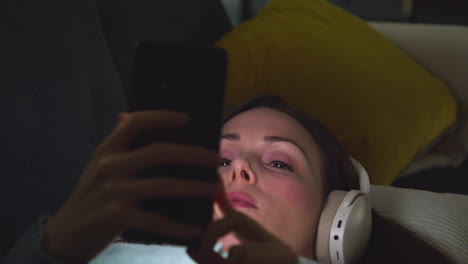 Woman Wearing Wireless Headphones Lying On Sofa At Home At Night Streaming Music Or Watching Movie On Mobile Phone 2