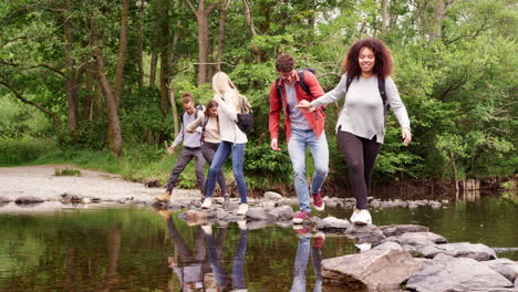 five young adult friends hold hands and help each other while crossing a stream and standing on stones during a hike