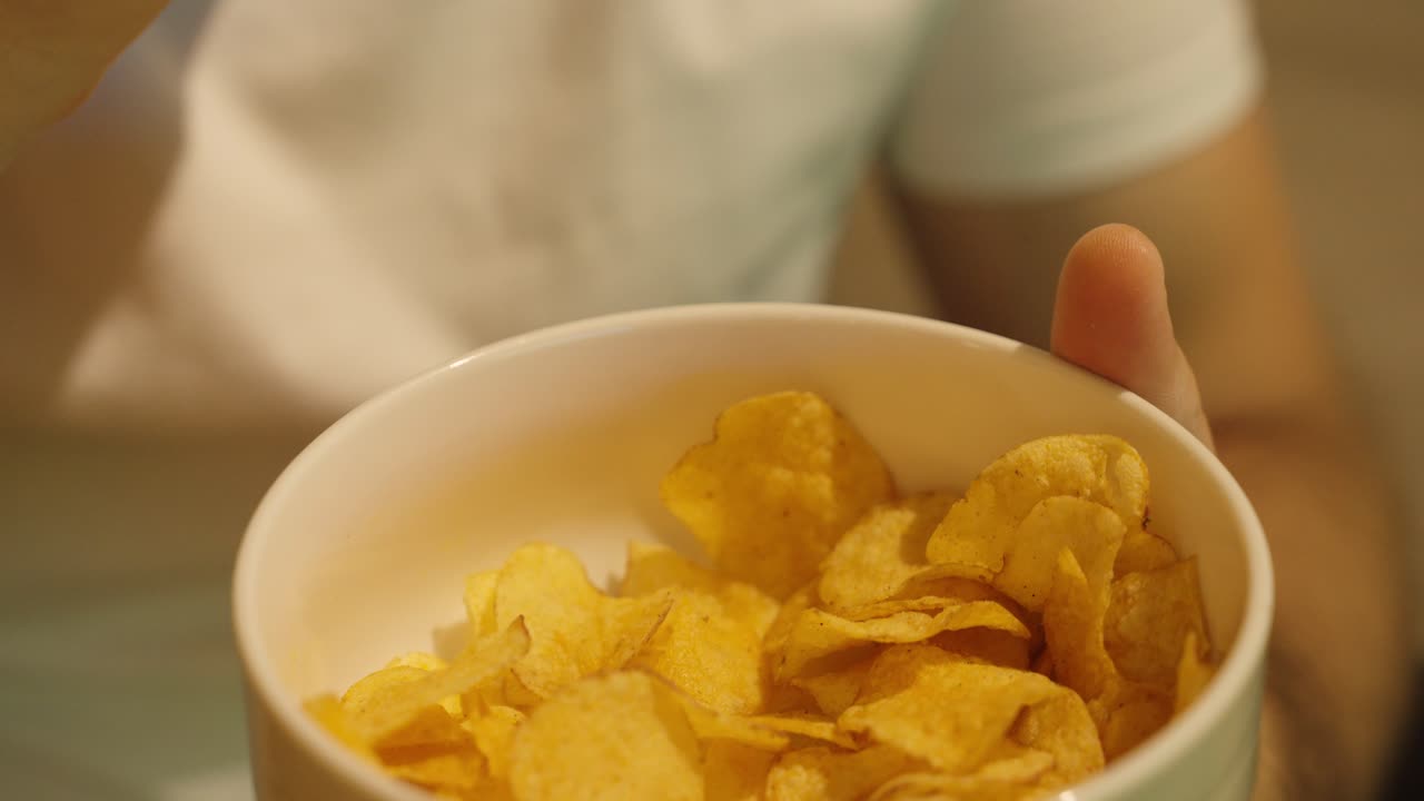 Primo piano di una persona che prende delle croccanti patatine fritte da una ciotola. Un momento di snack informale a casa, che mostra relax e comfort food