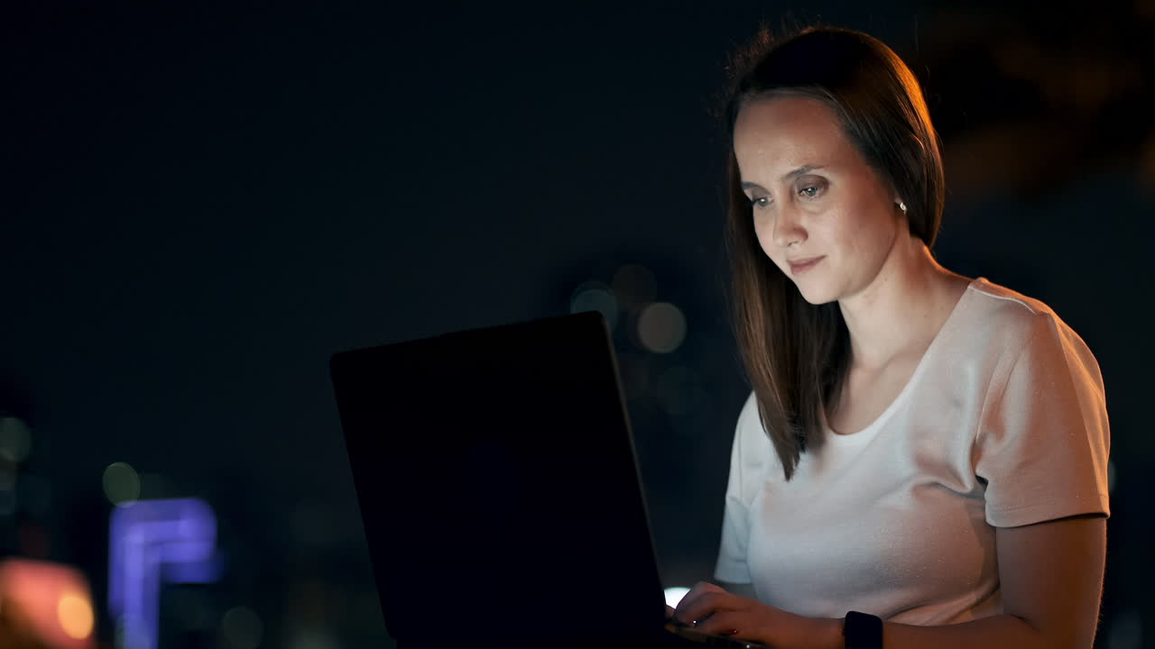 una mujer joven en una noche de verano sentada en la hierba en la ciudad mira la pantalla del portátil y escribe un mensaje en el teclado. programador freelance. trabajo nocturno remoto