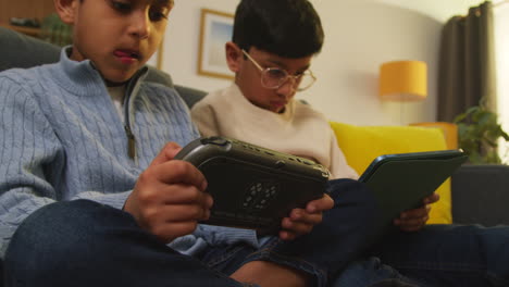 Two-Young-Boys-Sitting-On-Sofa-At-Home-Playing-Games-Or-Streaming-Onto-Digital-Tablet-And-Handheld-Gaming-Device-2