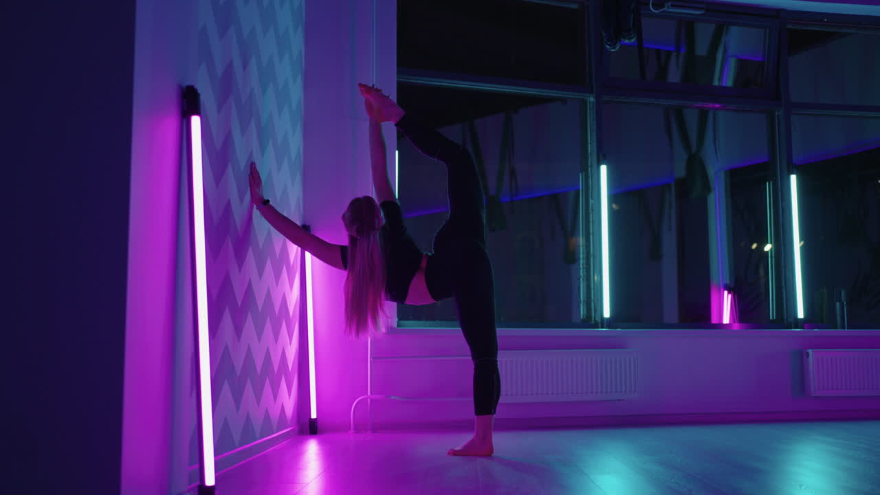 молодая гимнастка растягивается в фитнес-зале, опираясь на стену и поднимая ногу.