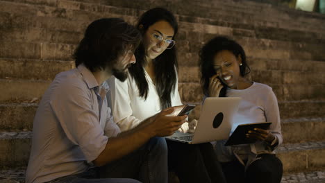 tres jóvenes sentados en las escaleras con dispositivos electrónicos por la noche en un día ventoso