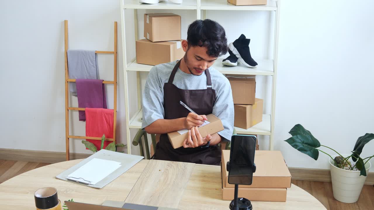 Young Asian Man In Apron Writing On Cardboard Box Preparing Package For Delivery