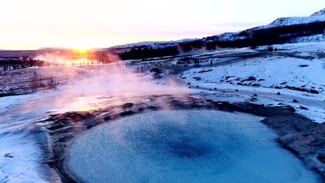 panoramic view of a geyser in geysir, iceland. smoke is getting out of the geyser with people walking all around the geyser, under the red and orange sunset.