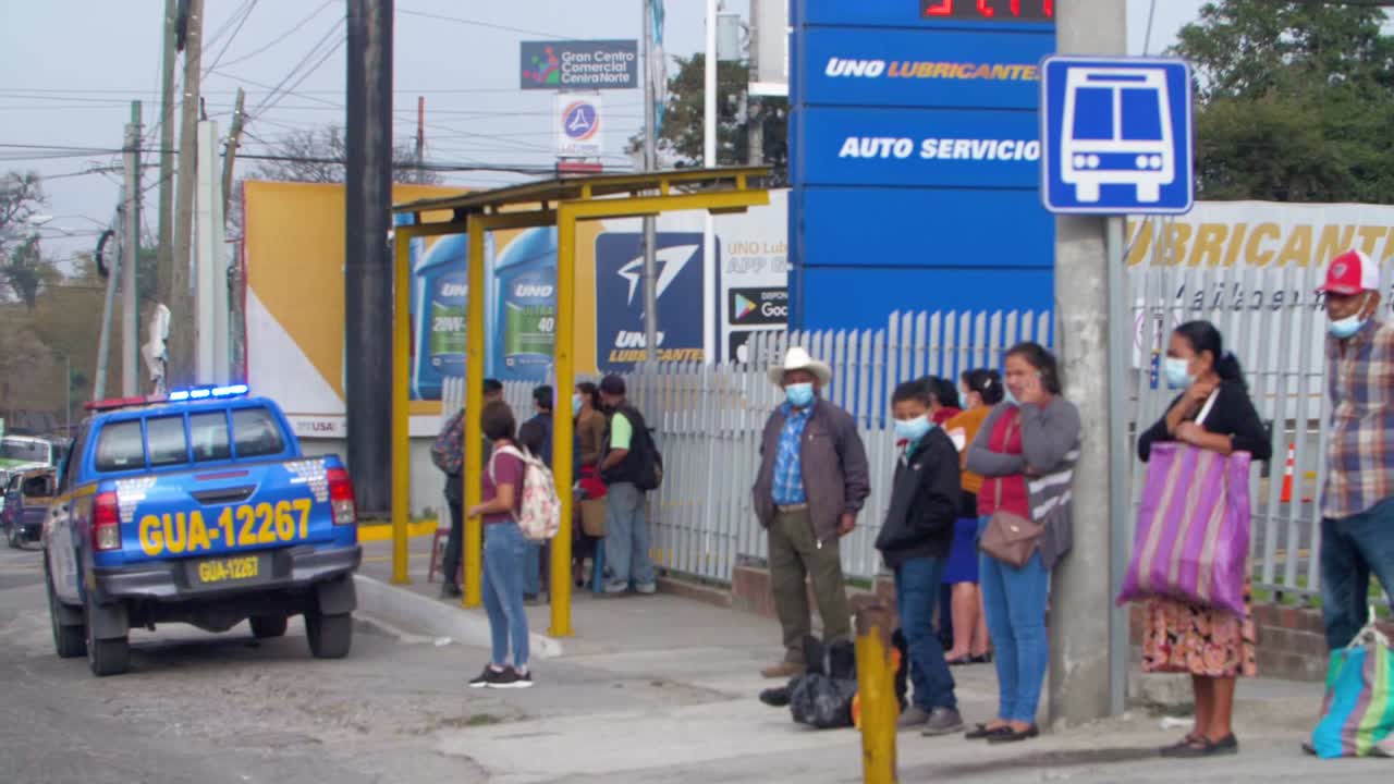 Slow motion camera moving left forward, people at bus stop wearing masks, police truck lights, Zone 18 Guatemala City, Guatemala