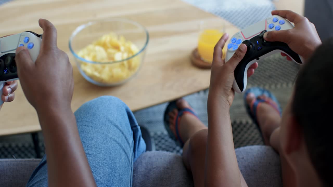 Second child entering, two kids pressing lit controllers and playing on couch with chips, juice