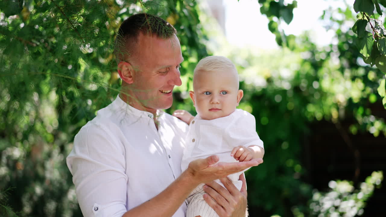 Happy Caucasian dad holding a baby stands under the tree. Smiling father talks to his son touching a tree branch. Man dances with his kid.