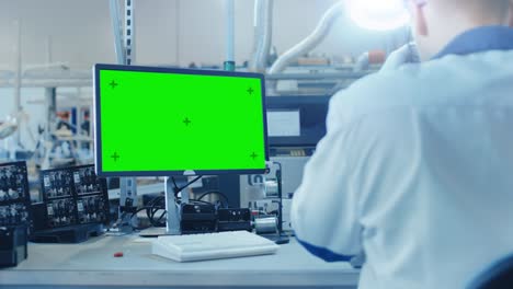 un trabajador de una fábrica de electrónica con abrigo de trabajo blanco inspecciona una placa de circuito impreso en una pantalla verde que está conectada a un microscopio digital.