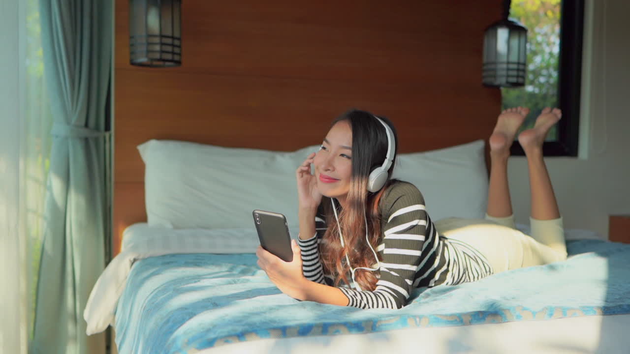 Beautiful Asian woman listening to music in headphones on her bed, happy time song and good morning concept