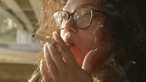 Black-woman-with-curly-hair-smoking-a-sigaret-underneath-a-bridge-during-sunset-in-autumn