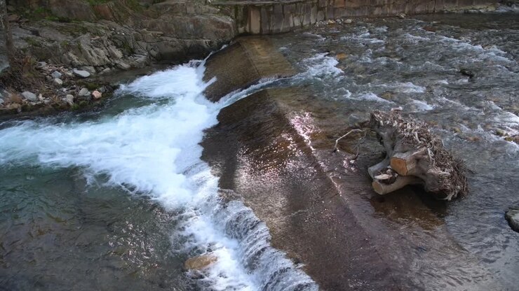 Acqua che scorre su un piccolo sbarramento fluviale creando una cascata in un paesaggio rurale naturale. Letto del fiume roccioso e corrente in movimento in un ambiente sereno.
