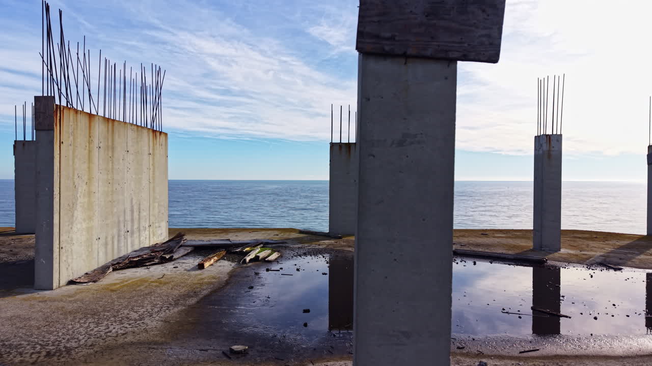 Old concrete pillars stand against a serene ocean backdrop as reflections dance in puddles under a bright clear sky