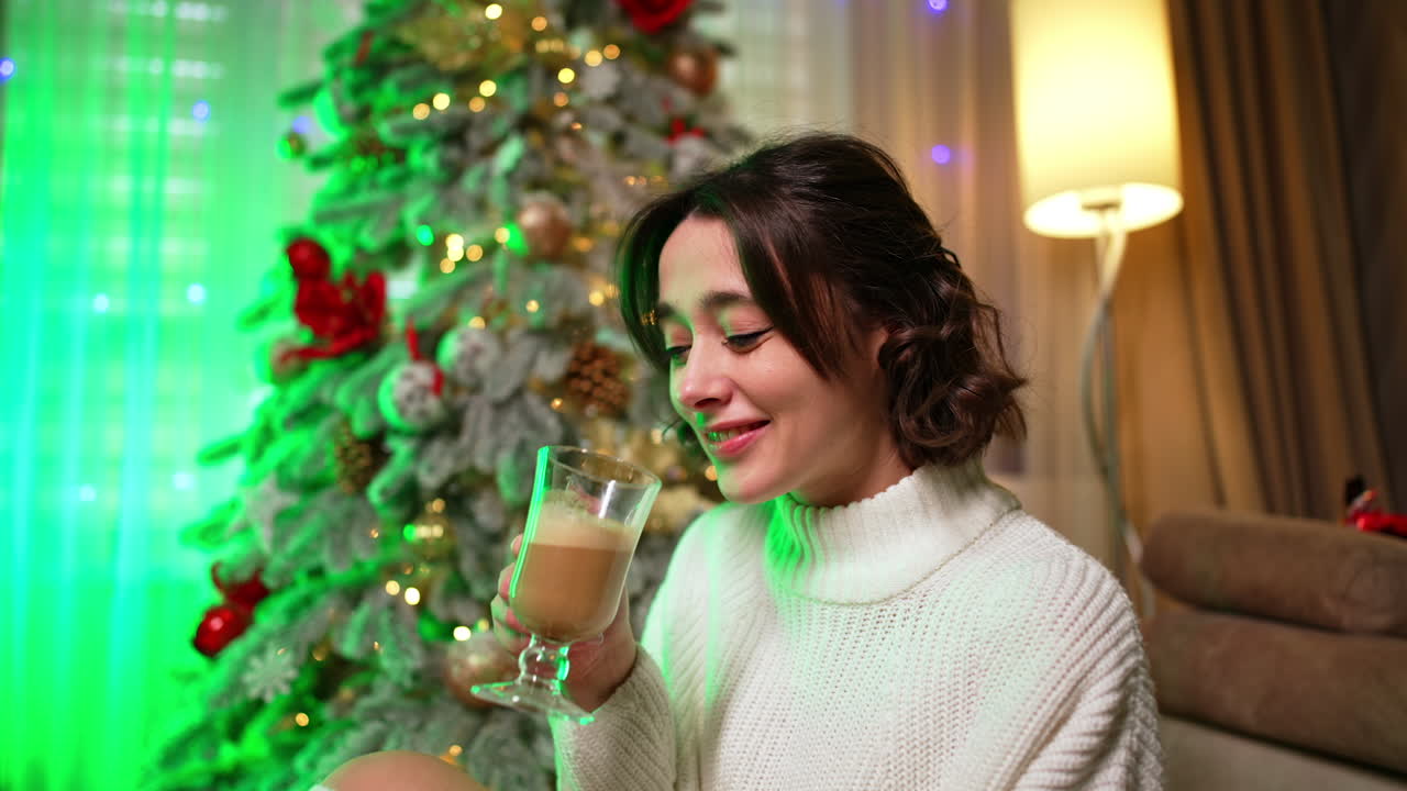 Approaching a beautiful girl in white sweater sits on the floor. Happy relaxed lady enjoys coffee near Christmas tree.