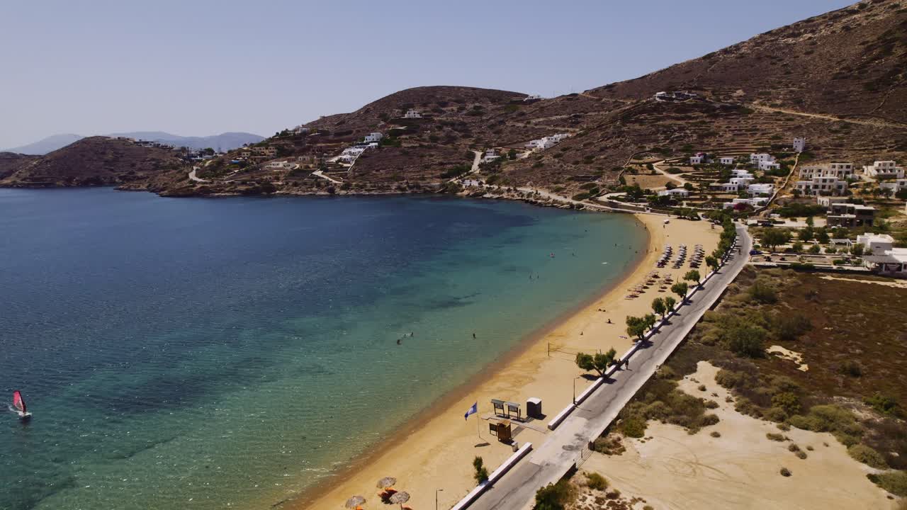Aerial drone view of a serene Greek island beach with clear turquoise waters, a coastal road, and traditional white houses nestled into the surrounding mountains