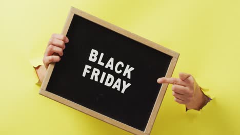 Hands-holding-and-pointing-at-black-board-with-black-friday-sale-text-in-white,-on-yellow-background