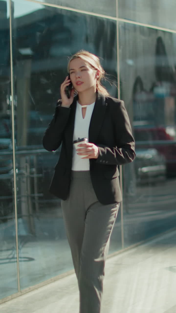 Young elegant banker woman walking confidently with coffee in hand while on phone call near modern glass building, exuding professionalism and focus as she strolls through bright urban environment