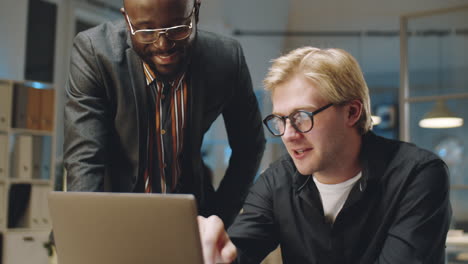 cheerful diverse colleagues discussing project on laptop in office at night