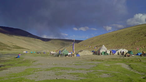 People,-hikers-walking-around-a-tent-camp-in-the-Himalayas,-base-camp-with-a-tall-prayer-flag-pole-at-Nimaling