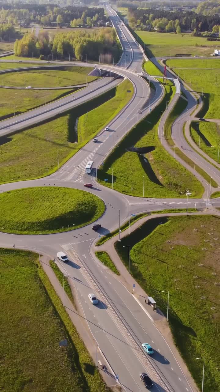 Katlakalns road interchange with rondeau, curved ramps, moving vehicles, green spring fields, complex road geometry, and forest line under clear sky and afternoon light, vertical aerial view