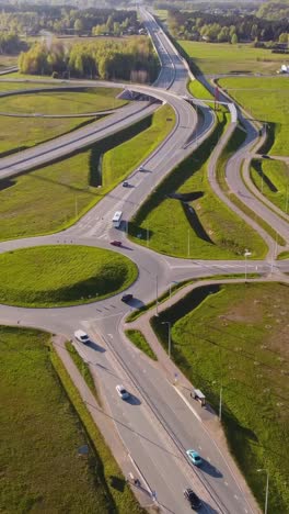 katlakalns road interchange with rondeau, curved ramps, moving vehicles, green spring fields, complex road geometry, and forest line under clear sky and afternoon light, vertical aerial view
