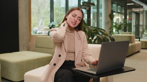 Tired young businesswoman suffers from spine neck muscle pain working on laptop in office lobby