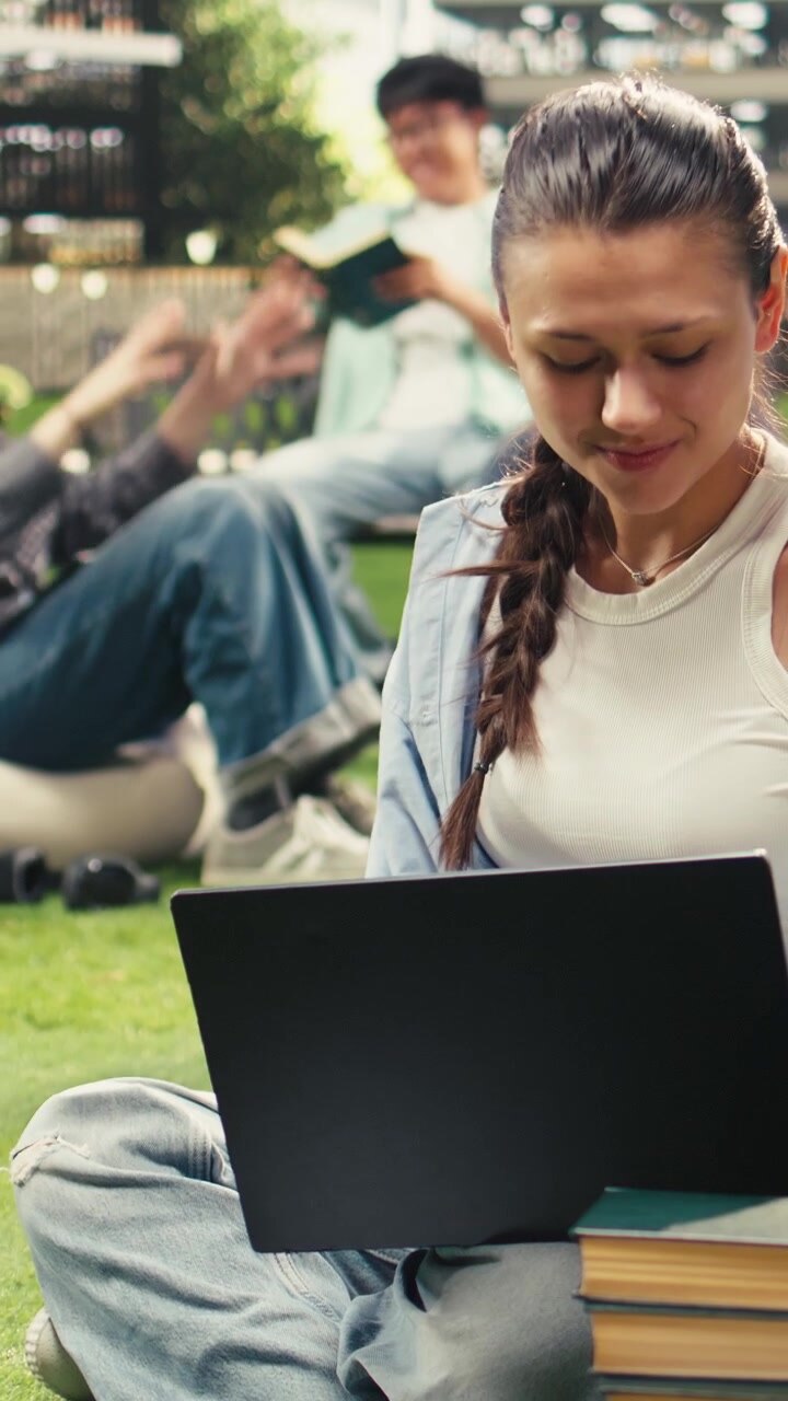 Video verticale: giovane donna sorridente che lavora al laptop seduta sull'erba in un campus moderno
