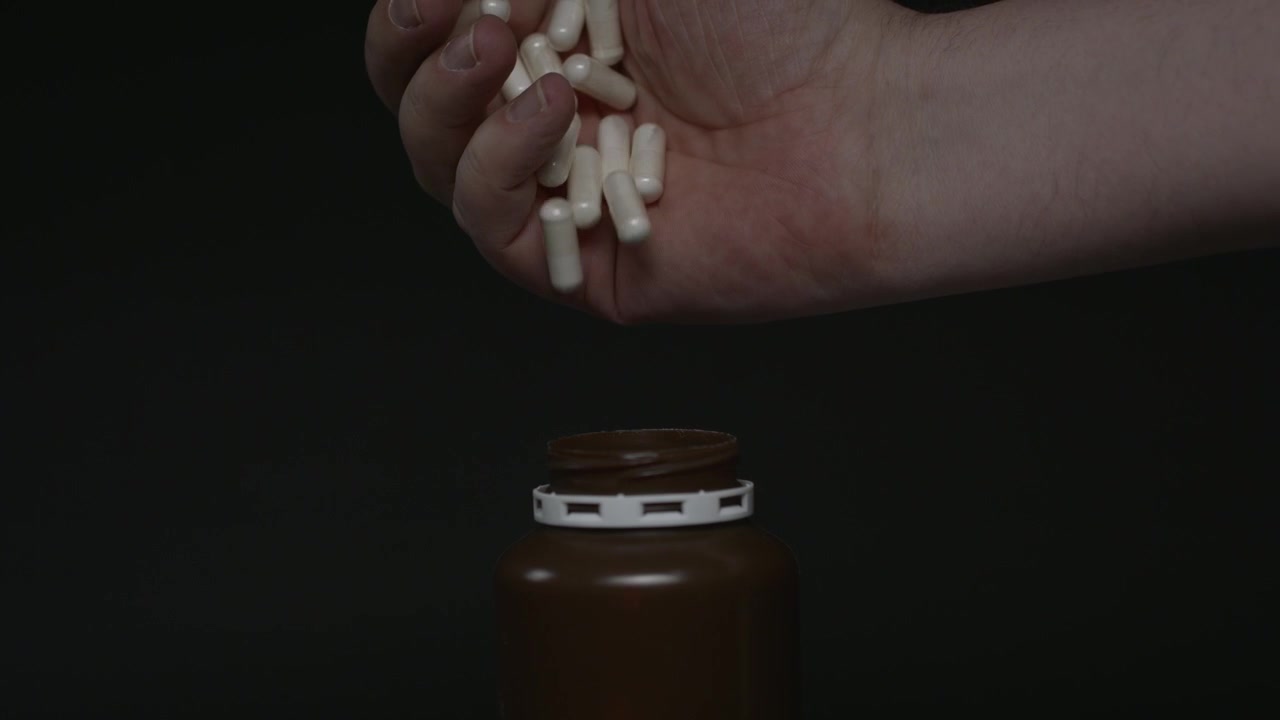 Close-up of a hand filling a brown medicine bottle with capsules in slow motion. Some pills fall outside the container on a black background.