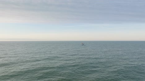 Aerial-view-of-a-coastal-fishing-boat-sailing-in-the-calm-Baltic-sea,-fisherman-dropping-fishing-net-in-the-water,-sunny-day,-distant-white-sand-beach,-wide-drone-shot-moving-forward