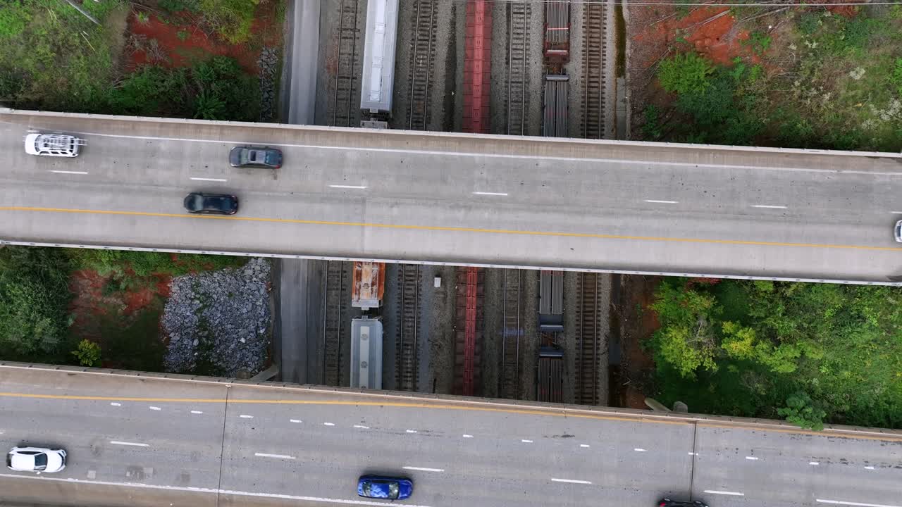 Aerial top down of traffic on overpass highway bridge with stopping train below. Slow forward Birds Eye in green suburb of USA.