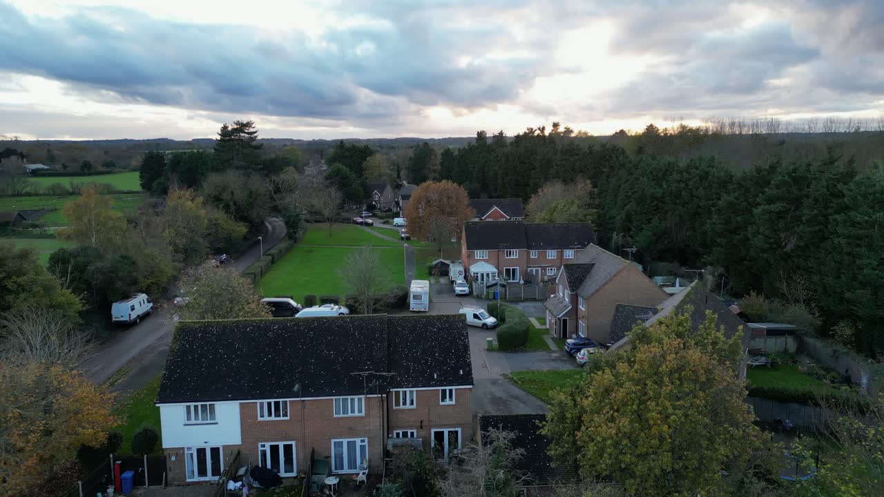 Low-altitude drone shot of a suburban UK neighbourhood. Houses, greenery, and vehicles are seen under a dramatic cloudy evening sky.
