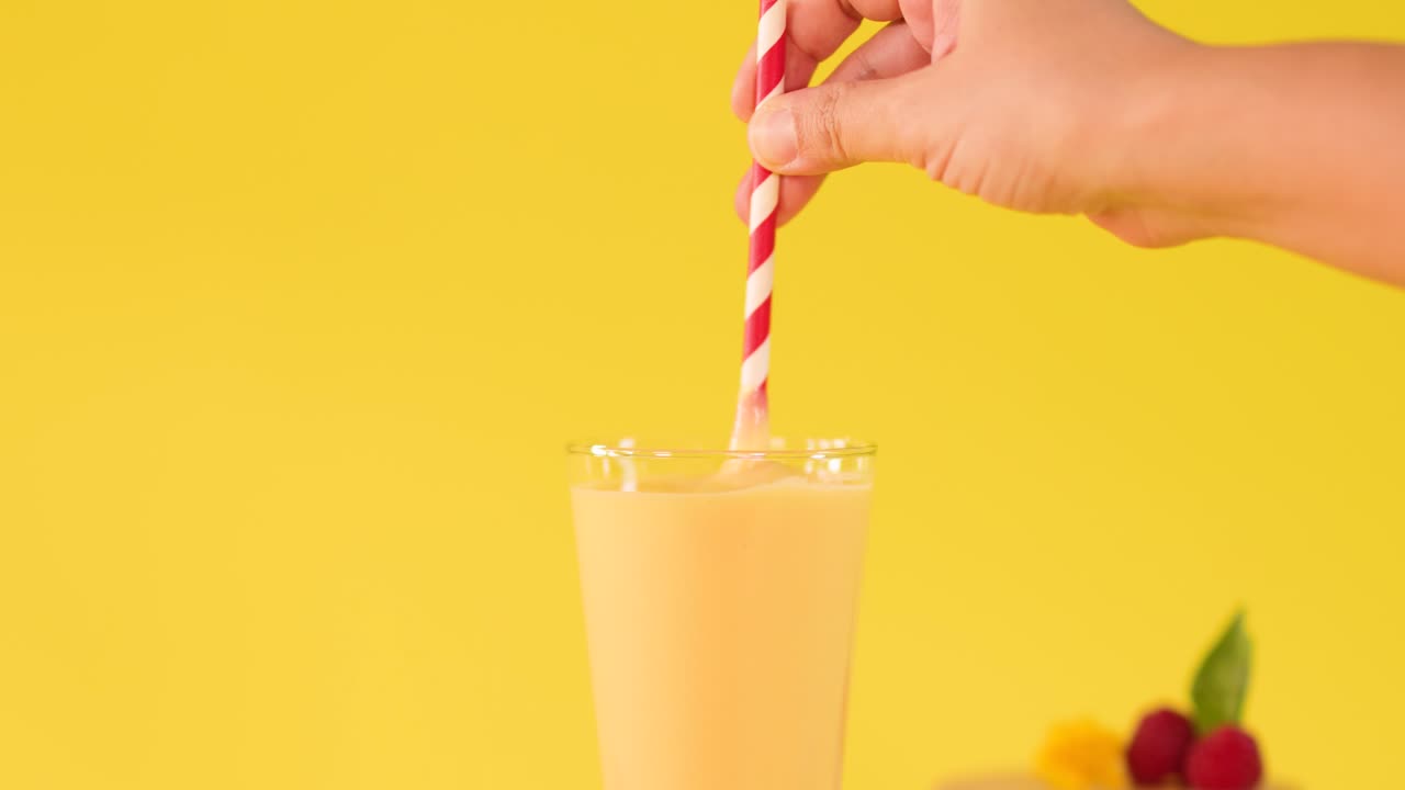 Una mano umana mescola un frullato freddo al mango con una cannuccia a righe su uno sfondo giallo brillante da studio