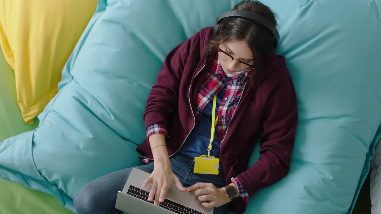 vista superior mujer feliz estudiante usando computadora portátil digital navegando en línea mensajes de redes sociales escribiendo un proyecto creativo relajándose en almohadas coloridas escuchando música en una oficina moderna