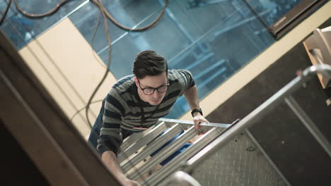 overhead perspective of young male worker wearing striped sweater and glasses climbing metal ladder inside modern industrial facility, showing effort, progress, safety, diligence, upward ambition