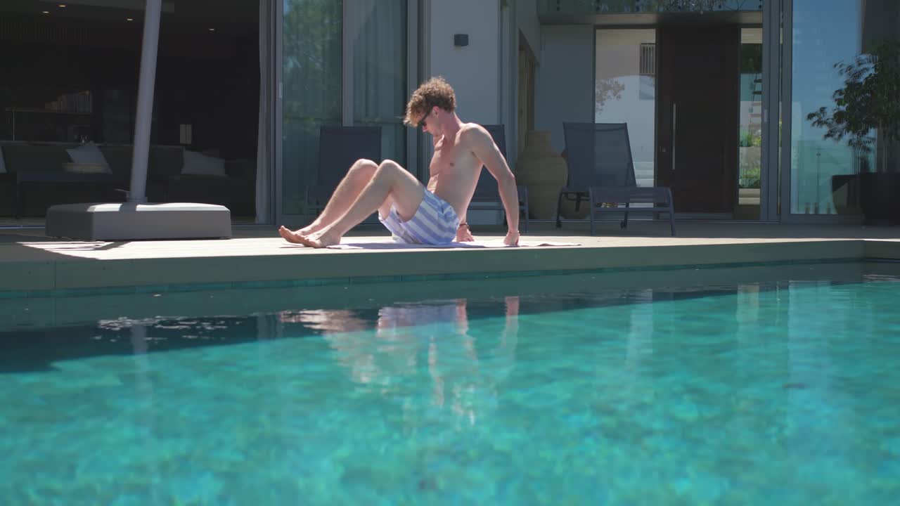 Die Kamera fährt zurück und zeigt einen Mann ohne Hemd, der seinen Oberkörper senkt, seine Sonnenbrille zurechtlegt und am Pool entspannt.