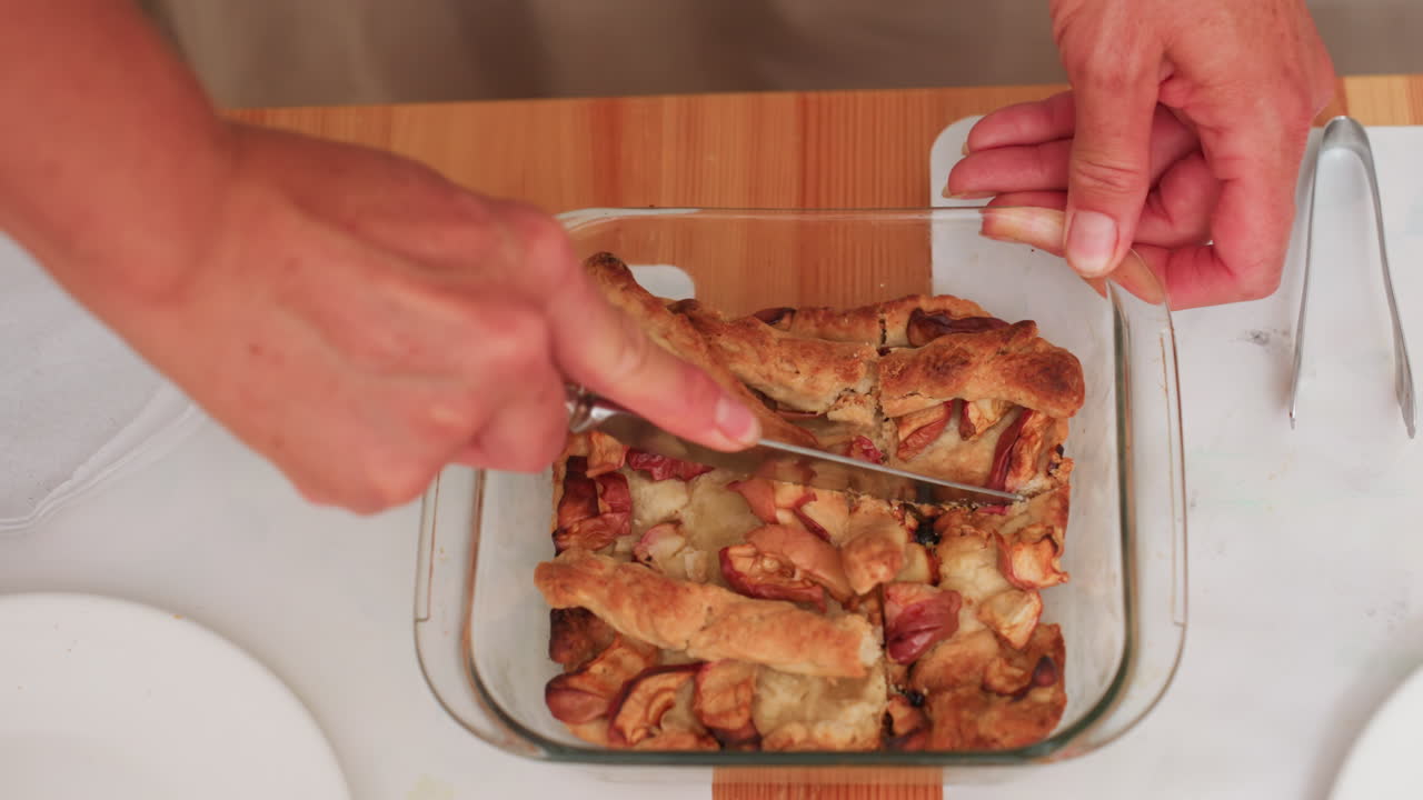 Vista de una mano de mujer cortando una tarta de manzana en una fuente de cristal sobre una mesa de madera, preparando raciones para los niños, un cuchillo cortando la masa con trozos de fruta horneada, servilletas y platos cerca.