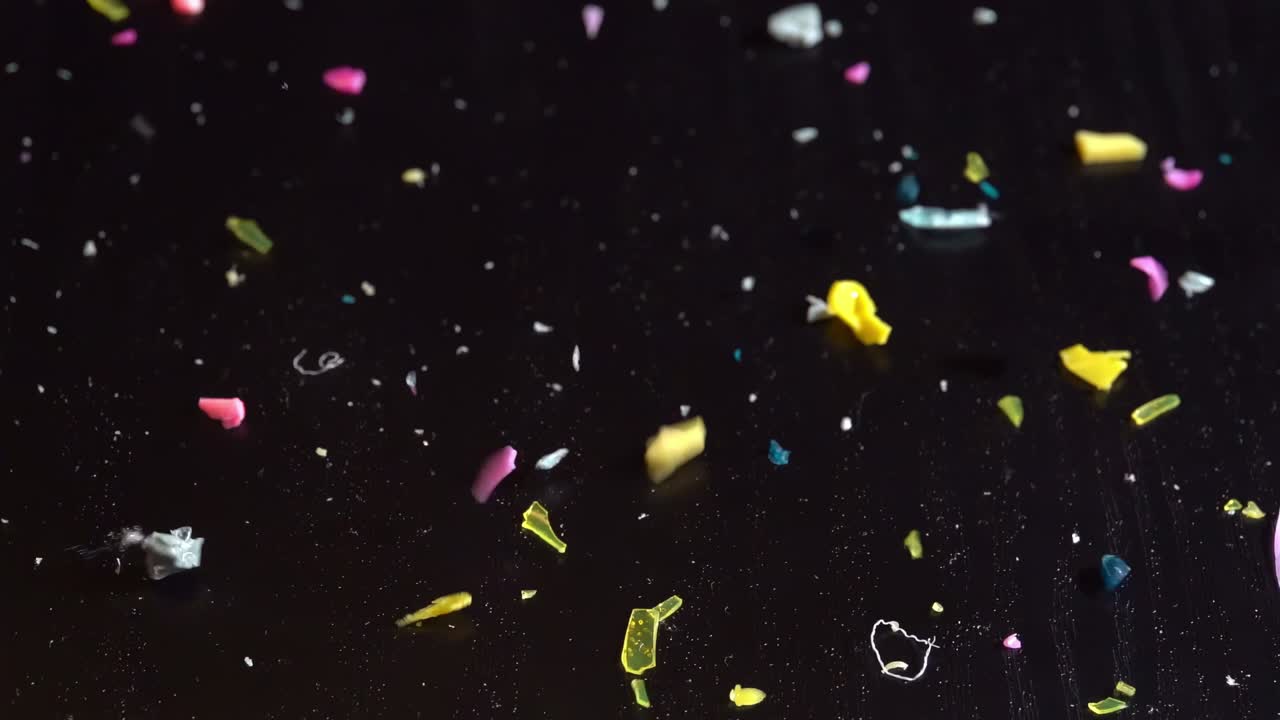 미세 플라스틱 조각들이 어두운 테이블 위에 느린 동작으로 반사되어 과도한 사용과 오염을 상징합니다.