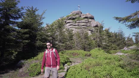 Cinematic-shot-of-young-male-tourist-walking-on-path-with-wooden-cross-at-the-top-of-a-hill-in-the-background,-front-view,-sliding-right