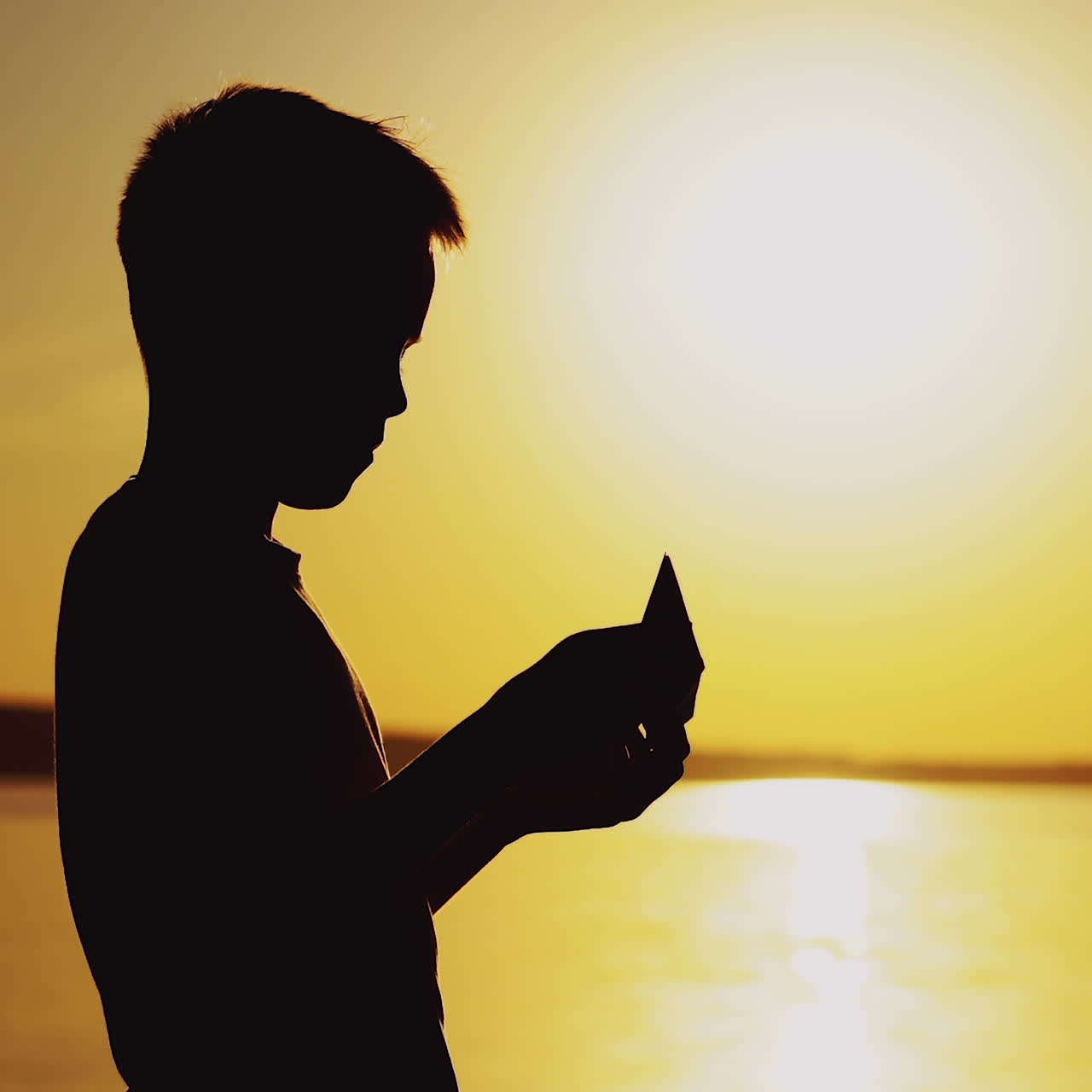 Young boy is making a paper ship standing near the river at the evening sunset. Silhouette of the boy playing with paper boat outside