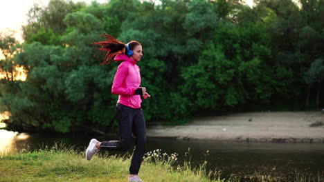 brunette with long hair in headphones runs along the river in the park in the morning at sunrise in the summer in a pink jacket and black pants