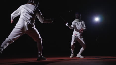 two fencers engage in a competitive match, one lunges forward with their sword while the other defends, the action is captured in a dark setting illuminated by a spotlight.