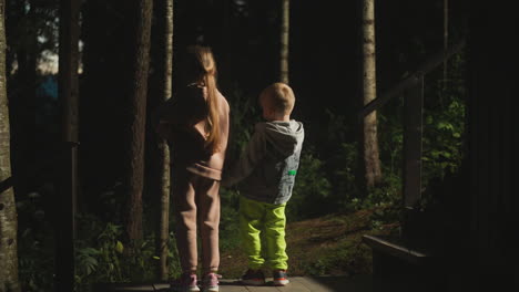 children on ground among wild dark forest. little girl and boy stand together on hotel pouch against night woods. kids look trees at woodland camp