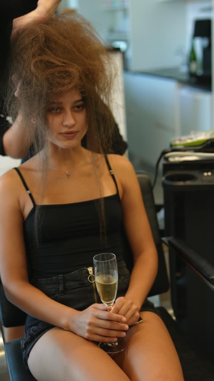 Giovane donna con capelli spettinati che tiene un bicchiere di champagne mentre si fa acconciare